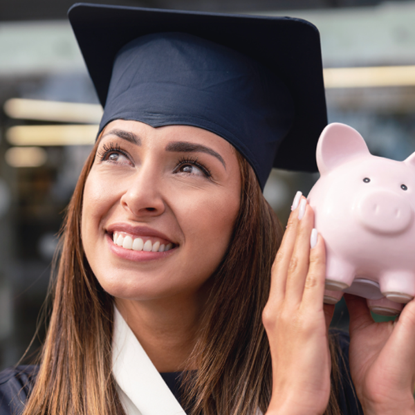 Woman wearing cap and gown holding piggy bank after saving and graduating college