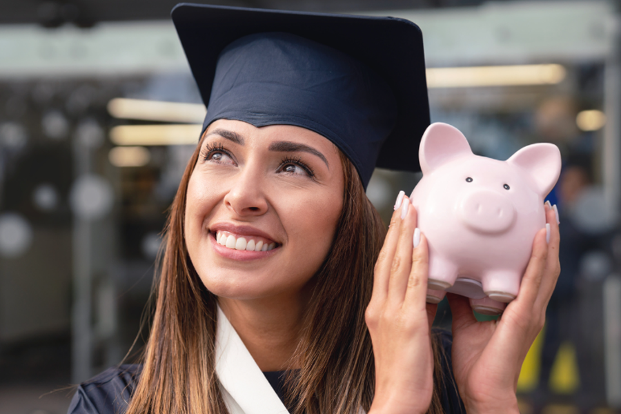 Woman wearing cap and gown holding piggy bank after saving and graduating college
